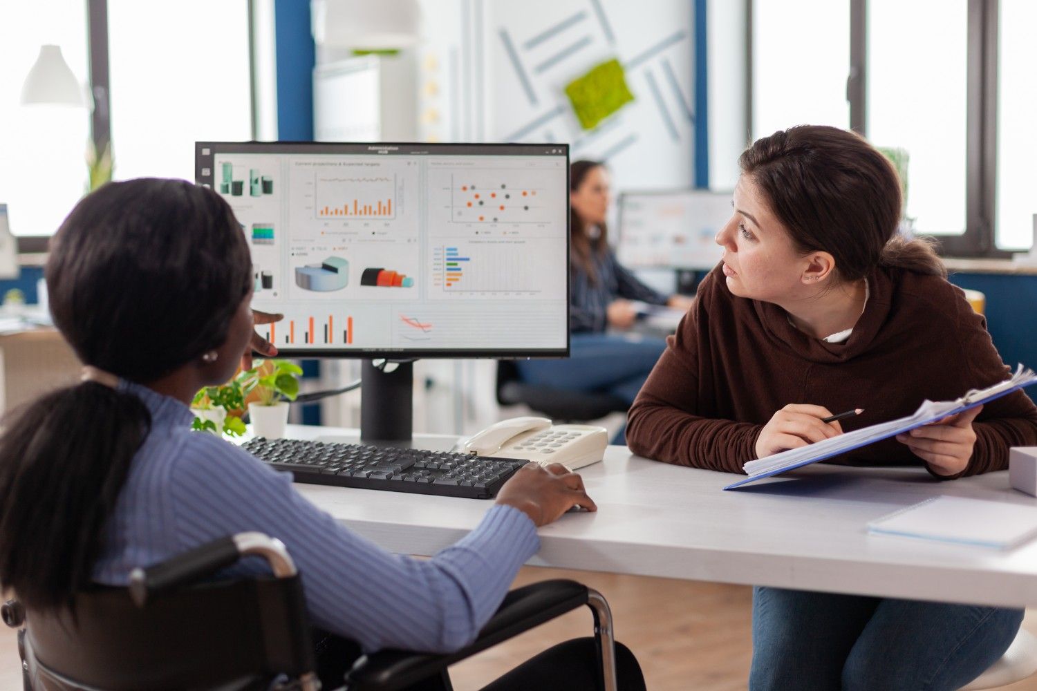 Two women sit at a desk in an office, reviewing graphs and charts displayed on a computer monitor.